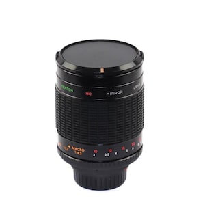 May include: A black Denton MC Mirror Lens with a macro setting. The lens has a ridged body with orange and green text. The lens cap is black and covers the top of the lens.