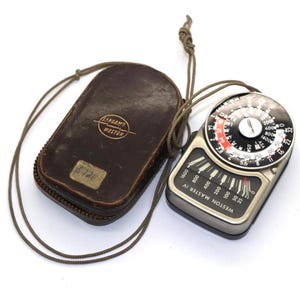 May include: A vintage Weston Master IV light meter with a leather case. The meter has a black and silver face with a needle and dial. The case is brown with the brand name and a serial number.
