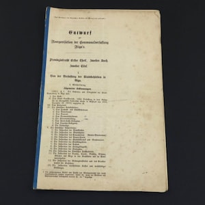 Könnte beinhalten: Ein altes Dokument mit deutschem Text und blauem Rücken. Der Titel "Entwurf" ist oben sichtbar, mit weiterem Text, der "Reorganisation der Sommanalverfallung Riga's" detailliert. Das Papier hat ein abgenutztes, antikes Aussehen.