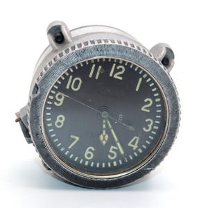 Può includere: Un orologio rotondo vintage con quadrante nero e numeri bianchi. L'orologio ha un involucro in metallo color argento con due piccole alette di montaggio. Le lancette sono bianche e il quadrante ha una piccola lancetta dei secondi. L'orologio è probabilmente uno strumento aeronautico o automobilistico.