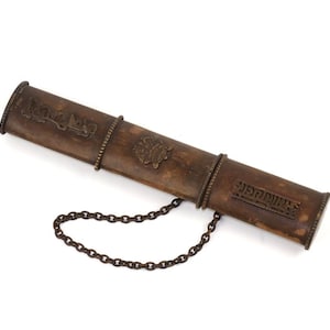 Puede incluir: Un estuche cilíndrico marrón antiguo con bandas y adornos metálicos decorativos. El estuche tiene una cadena y parece estar hecho de madera o un material similar. El estuche tiene texto.