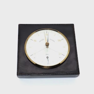 May include: A square black leather-covered barometer featuring a circular white dial with gold-toned hands and a gold-toned metal rim. The dial displays the German text "WEGEN" and "SCHON" along with numerical markings and a small secondary dial.
