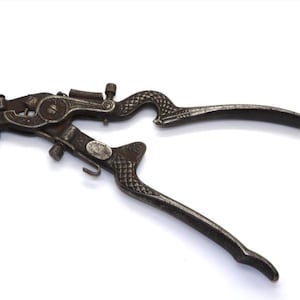 May include: An antique metal saw set tool with ornate, textured handles. The tool is dark grey with a spring and various levers and pins. The handles have a decorative, scale-like pattern. The tool is designed to adjust the set of saw teeth.