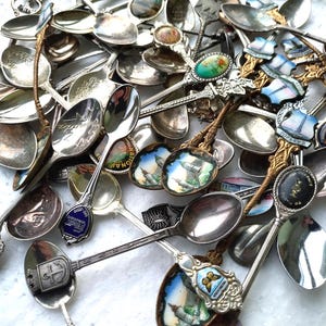 Könnte beinhalten: Eine Sammlung von Vintage-Silberlöffeln mit verzierten Griffen und dekorativen Schalen. Einige Löffel weisen Emaille-Designs mit farbenfrohen Landschaften und Wahrzeichen auf. Die Löffel variieren in Größe und Stil und zeigen eine Vielzahl von Details.