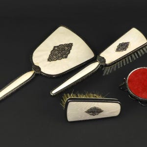 May include: A vintage vanity set featuring a hand mirror, two brushes, and a small round container with a red cushion. The set has a cream-colored finish with ornate silver-toned trim and dark accents.