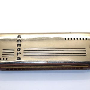 May include: A vintage Sonora harmonica with a polished silver cover and red trim. The word "sonora" is printed vertically. The harmonica features a series of holes and lines. A classic musical instrument, likely from the mid-20th century.