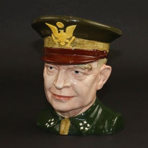 May include: A ceramic bust of a military figure, with a green uniform and cap adorned with a gold emblem. The bust has a detailed face with painted features, including a red mouth and blue eyes. A collectible item.