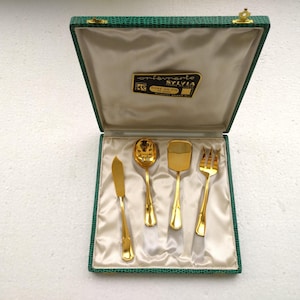 May include: A vintage gold-toned flatware set in a green faux-leather box. The set includes a knife, spoon, spatula, and fork, all displayed on a satin lining. The box has a gold clasp and a label that reads "Fontanarte Sylvia".