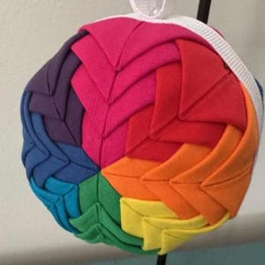 May include: A colorful, round fabric ornament with a rainbow pattern. The ornament features a series of folded fabric segments in shades of red, orange, yellow, green, blue, purple, and pink. A white ribbon is attached for hanging.