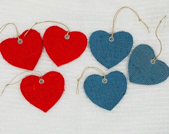 Puede incluir: Seis etiquetas de yute en forma de corazón en rojo y azul. Cada etiqueta tiene un ojal metálico y un lazo de cuerda natural para colgar. Las etiquetas están dispuestas sobre una superficie blanca, ideales para manualidades o decoraciones.