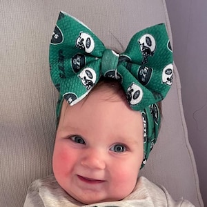 May include: A baby wearing a green and white bow headband and a gray sweatshirt with a football and the word "GAME" in green. The baby is sitting in a chair and smiling.