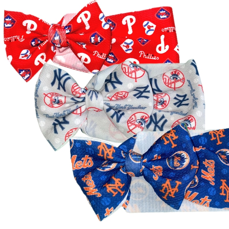 May include: Three fabric hair bows with different baseball team logos. The first bow is red with white and blue accents and the Philadelphia Phillies logo. The second bow is white with blue accents and the New York Yankees logo. The third bow is blue with orange accents and the New York Mets logo.