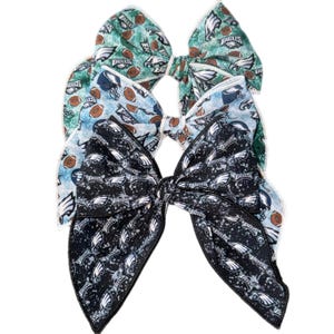 May include: Three large fabric hair bows in a layered arrangement. The bows feature the Philadelphia Eagles logo and football-themed patterns in green, blue, and black. Each bow has a white stitched edge.