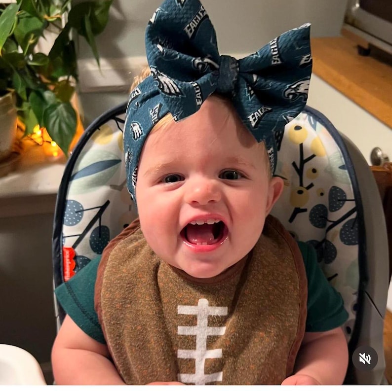 May include: A baby wearing a green shirt and a brown bib with a football design. The baby is wearing a teal headband with a white "Eagles" logo.