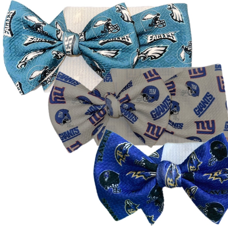 May include: Three fabric hair bows featuring NFL team logos. One is light blue with Eagles logos, another is gray with Giants logos, and the third is blue with Ravens logos. Each bow has a matching fabric band.