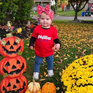 May include: A young child wearing a red shirt with the words "Phillies WS * 2022" printed on it. The child is standing in front of a stack of jack-o-lanterns and a patch of yellow mums.