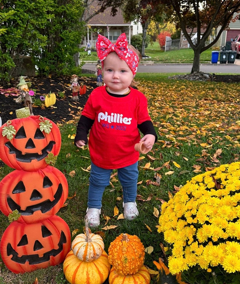 May include: A young child wearing a red shirt with the words "Phillies WS * 2022" printed on it. The child is standing in front of a stack of jack-o-lanterns and a patch of yellow mums.