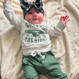 May include: A baby wearing a white long-sleeved shirt with a green eagle graphic and the text "Sundays Are For The Birds". The baby is also wearing green pants and a black and green bow headband.