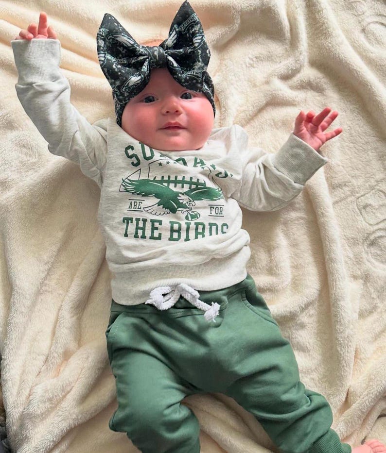 May include: A baby wearing a white long-sleeved shirt with a green eagle graphic and the text "Sundays Are For The Birds". The baby is also wearing green pants and a black and green bow headband.