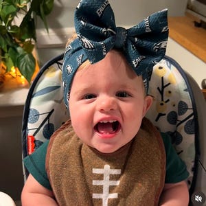 May include: A smiling baby wearing a dark green and white Philadelphia Eagles bow headband and a brown football-themed bib. The bib has a white football design. The baby is sitting in a high chair.