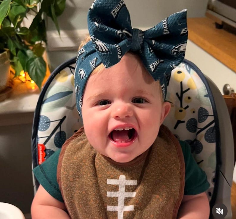 May include: A smiling baby wearing a dark green and white Philadelphia Eagles bow headband and a brown football-themed bib. The bib has a white football design. The baby is sitting in a high chair.