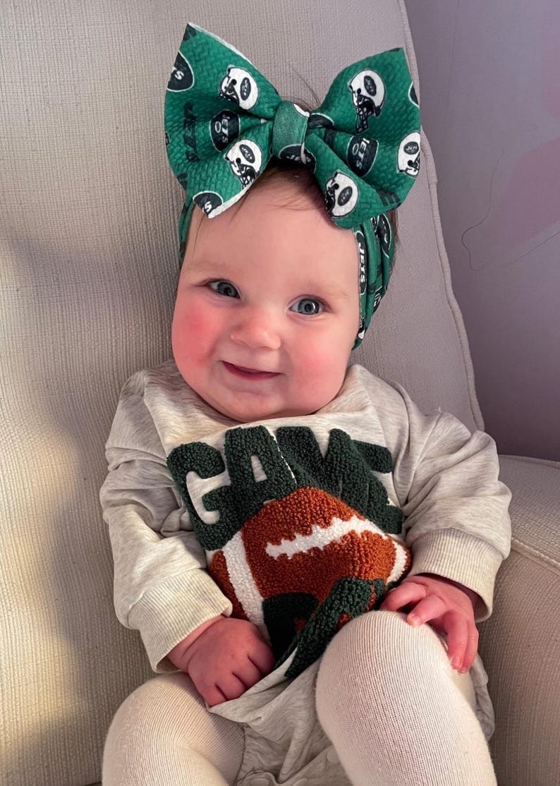 May include: A baby wearing a green and white bow headband and a gray sweatshirt with a football and the word "GAME" in green. The baby is sitting in a chair and smiling.