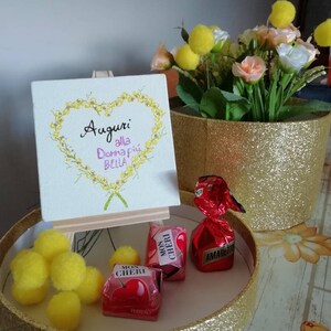Può includere: Una scatola regalo dorata con pompon  gialli e cioccolatini avvolti. Un piccolo cavalletto sostiene una tela bianca con il testo "Auguri alla donna più bella" in lettere nere e rosa.