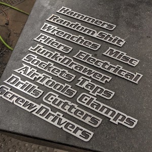 May include: A set of metal labels with tool names, including "Hammers", "Random Shit", "Wrenches", "Pliers", "Electrical", "Junk Drawer", "Sockets", "Taps", "Air Tools", "Clamps", "Drills", "Cutters", and "Screw Drivers".
