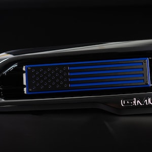 May include: A black GMC truck grille with a blue and black American flag emblem. The flag has a thin blue line across the stripes.