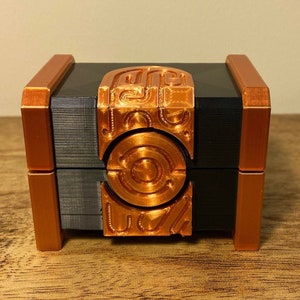 May include: A small, black and copper-colored box with intricate geometric designs. The box has a hinged lid and a decorative front panel with circular and linear patterns. The box is sitting on a wooden surface.