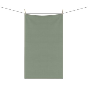May include: A sage green rectangular towel hanging from a clothesline with wooden clothespins. The towel is made of a soft fabric and has a simple, minimalist design. The background is plain white.