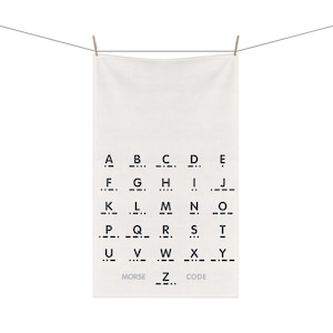 May include: White towel with black Morse code alphabet printed on it. The towel is hanging from a clothesline with wooden clothespins. The words "MORSE CODE" are printed at the bottom.