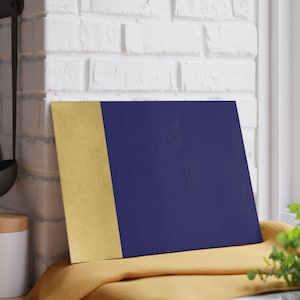 May include: A square, two-toned decorative item. The left side is gold, and the right side is navy blue. It rests on a yellow surface, with a white brick wall in the background. The item is leaning against a window.