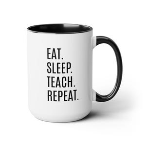 May include: White ceramic mug with a black interior, handle, and rim. The mug features the text "EAT. SLEEP. TEACH. REPEAT." in a bold, black font. The mug is designed for hot drinks.