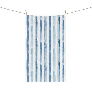 May include: A white towel featuring vertical stripes in shades of blue, reminiscent of watercolour strokes. The towel is suspended from a washing line with wooden pegs. The design is simple and contemporary.
