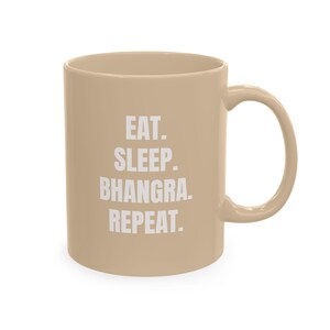 May include: A light tan ceramic mug featuring the white text EAT. SLEEP. BHANGRA. REPEAT. printed in a bold, sans-serif font on the front. The mug has a smooth, cylindrical body and a curved handle on the right side for a comfortable grip.