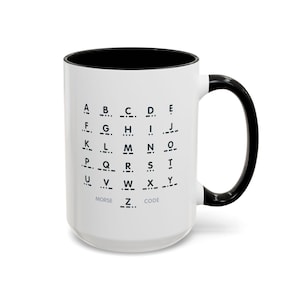 Könnte beinhalten: Weißer Keramikbecher mit schwarzem Innenraum und Henkel. Der Becher zeigt das Morsealphabet in Schwarz, darunter die Worte "MORSE CODE". Ein stilvoller Becher für Kaffee oder Tee.