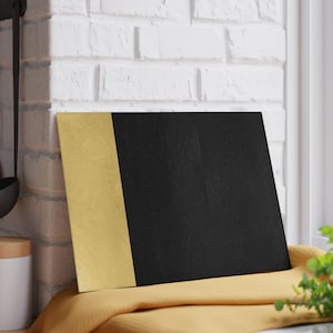 May include: A square chopping board with a black surface and a vertical gold-coloured accent on the left side. The board is resting on a yellow cloth, with a white brick wall in the background.