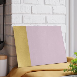 May include: A rectangular chopping board with a gold vertical stripe on the left and a light pink section on the right. The board is resting on a yellow cloth, with a white brick wall in the background.
