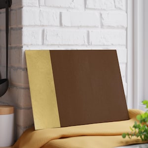 May include: A rectangular chopping board with a gold vertical stripe on the left and a brown surface on the right. The board rests on a yellow cloth near a window.