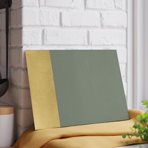 May include: A rectangular chopping board with a gold vertical stripe on the left and a muted green surface on the right. The board rests on a yellow cloth. The background features a white brick wall.