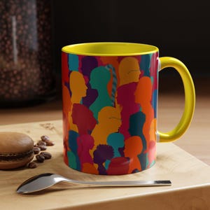 Puede incluir: Una taza de cerámica con asa e interior amarillos. El exterior presenta un colorido patrón de diversas siluetas humanas en tonos naranja, verde azulado, morado y rojo. La taza está sobre una superficie de madera con una cuchara y un pastel.