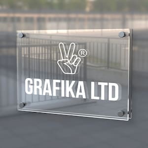 Puede incluir: Un letrero de acrílico transparente con el texto "GRAFIKA LTD" en blanco, junto con un símbolo de mano. El letrero está montado con herrajes plateados. El fondo está borroso, sugiriendo un entorno exterior.