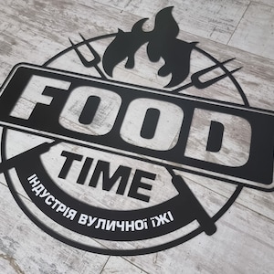 May include: Black metal sign with the words "FOOD TIME" in a bold, sans-serif font. The sign includes a circular design with stylized flames and forks, and text in a foreign language. The sign is placed on a light wood-grain surface.