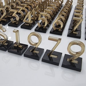 May include: A collection of gold-colored, transparent numbers and a dollar sign, each mounted on a small, black square base. The numbers are arranged in rows, with a variety of digits visible, suggesting a numerical or financial theme.