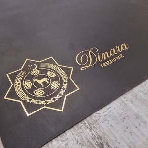 May include: A dark brown placemat with a gold emblem and text. The emblem is a star shape with a horse in the center. The text reads "Dinara restaurant" in a cursive font.