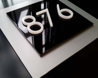 May include: A modern house number sign featuring the numbers 876 in white against a glossy black background. The sign is mounted on a silver-colored base, creating a sleek, contemporary look.
