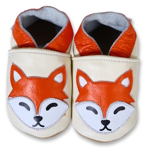 Puede incluir: Un par de zapatos de beb&eacute; de cuero color crema con un dise&ntilde;o de cara de zorro. El zorro tiene pelaje naranja, ojos blancos y una nariz negra.