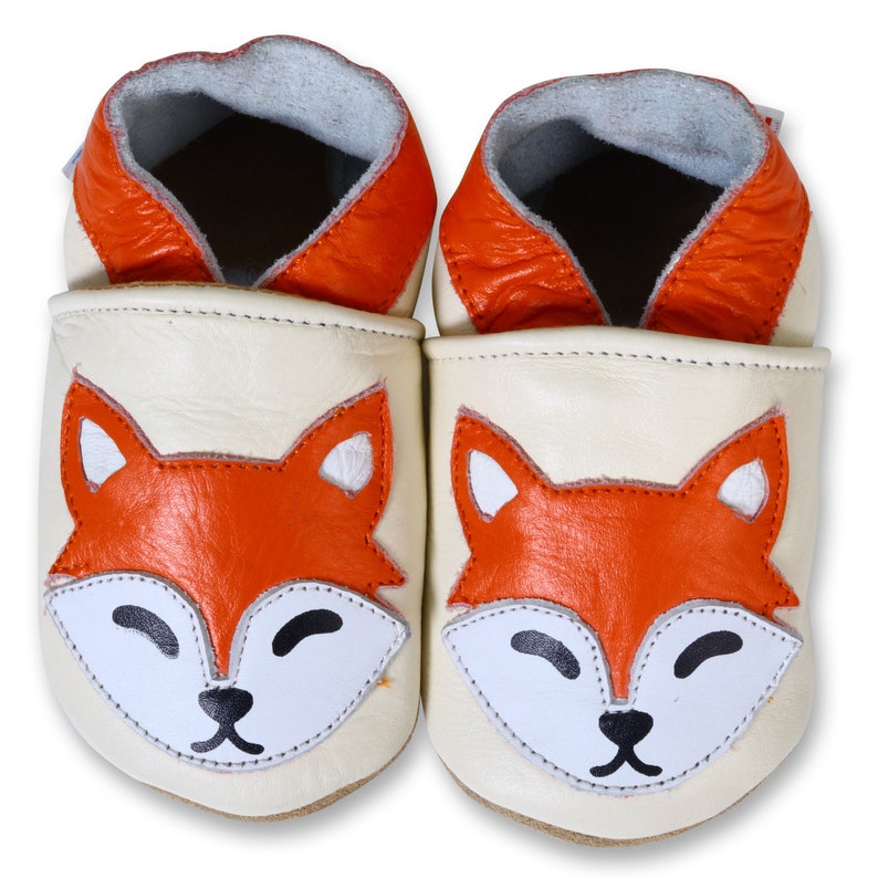 Puede incluir: Un par de zapatos de beb&eacute; de cuero color crema con un dise&ntilde;o de cara de zorro. El zorro tiene pelaje naranja, ojos blancos y una nariz negra.