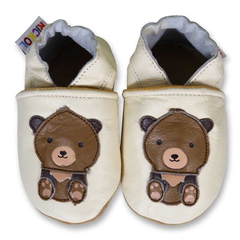 Puede incluir: Un par de zapatos de beb&eacute; de cuero color crema con un dise&ntilde;o de oso marr&oacute;n. Los zapatos tienen una suela suave y flexible, perfectos para gatear y caminar.